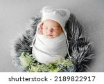 Small photo of Newborn baby child swaddled in white fabric sleeping. Sweet infant kid wearing hat napping on fur