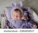 Small photo of Newborn baby girl wearing knitted hat with rabbit ears holding bunny toy and looking at camera. Infant child kid lying swaddled in purple fabric studio portrait