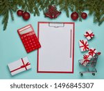 Shopping trolley with gift boxes, clipboard with blank sheet and fir tree branches  with decoration on green background. Top view. Christmas and New Year sale. Planning budget. Cost of holiday