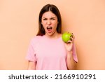 Young hispanic woman holding an apple isolated on beige background screaming very angry and aggressive.