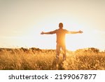 Feelings of hope, power and freedom in nature. Young man standing in a field with arms up feeling the rays on the warm sunshine light. Mind body spirit concept. 