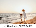 A young mother holds her little daughter by the hand and together they walk along the ocean towards the sunset. Girls in white dresses and with long hair that develops the wind