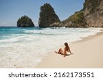 Young girl in a yellow swimsuit sits on a tropical white sand beach with turquoise water. Vacation trip to Diamond Beach in Nusa Penida Bali Indonesia