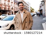 Young handsome smiling stylish man in trench coat joyfully walking through city street