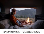 Woman enjoys bonfire, sitting and relaxing in the vehicle trunk with pillows. Traveling by car, romance at picnic in the mountains
