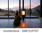 Small photo of Woman enjoy great view on mountains while sitting near fireplace at modern living room at sunset . Concept of rest in houses or cabins on nature. Solitude in nature and escape from everyday life