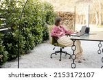 Young woman works on laptop at cozy outdoor workspace in the garden. Concept of remote work from comfortable home office or work during vacations