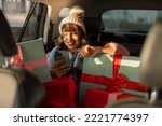 Young woman dressed for the holidays drives in car on back seat with beautifully wrapped Christmas presents and using phone. In anticipation of the winter holidays, preparation and shopping concept