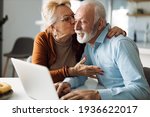 Small photo of Mature woman kissing her mature husband while he uses a computer at home