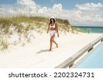 Small photo of Beautiful Hispanic woman in a white bikini with an American flag on whet sandy beach emerald waters of the emerald coast the Gulf Of Mexico with a hat and a drink and beach bag woman Pensacola beac
