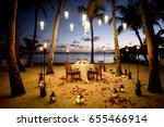 A Table Set up for a romantic meal on the beach with lanterns and chairs and flowers with palms and sky and sea in the background