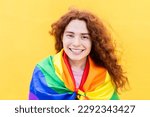 Portrait of red haired non binary man with rainbow flag smiling at camera over yellow background. LGBT community, diversity, transgender and non-binary people concept