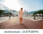 a bright beautiful girl in a light dress and hat walks along the streets of old town of Monaco in sunny weather, a residential area of principality and walking people in the background