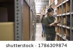 Small photo of e-shop female employee counting cardboard boxes on shelf and writing on clipboards in warehouse. beautiful women coworkers working together in stockroom doing stock taking. colleague wear uniform