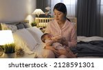Small photo of tired new mother sitting by lamp is dozing off while bottle feeding her newborn child at midnight in the bedchamber.