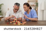 Small photo of caring asian woman care attendant giving instructions while assisting elderly patient go through rehab treatment for Parkinsonâ€™s disease with building blocks at home