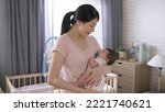 Small photo of asian first time mom is singing lullaby quietly to her baby girl while cradling her in the arms during naptime inside a bedchamber.