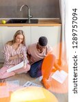Small photo of Young mixed-race couple of diverse students in casual wear, sitting together on kitchen floor at home working at their coursework having concentrated looks in lecture notes.