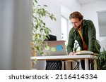 Bearded young confident guy stand behind office desk with laptop thinking working alone. handsome male in casual wear engaged in office work, planning strategy, deadlines. at workplace