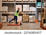 cheerful caucasian male staff dressed in working clothes and helmet in warehouse having fun standing on pallet truck jack, driving among shelves, enjoy free time, leisure at work place