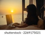 Woman sitting on bed under white blanket near window, looking to Beautiful sunset or sunrise above sea, reflection of sun in the water, surfing on internet on laptop, thoughts thinking and planning.