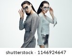 Portrait of two young females in silver outfit and mirrored sunglasses looking at camera. Female models in futuristic look standing on grey background.