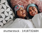 Small photo of Top view of two girls laughing with eyes closed lying on bed having a sleepover. Girls with fancy sleep masks over the head laughing and having fun during a sleepover.