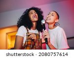 Best friends singing into a microphone on karaoke night. Two cheerful young women singing their favourite song at a house party. Happy female friends having a good time during the weekend.