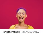 Small photo of Quirky and vibrant. Excited young woman sticking her tongue out with her eyes closed while standing against a pink background. Fashionable young woman wearing makeup with purple hair in a studio.