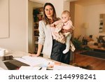 Creative businesswoman holding her baby while standing behind her desk. Working mom planning a new project in her home office. Female interior designer balancing work and motherhood.