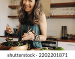 Smiling senior woman having a delicious buddha bowl in her kitchen. Excited woman serving herself some healthy vegan food at home. Mature woman taking care of her aging body with a plant-based diet.