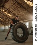 Small photo of Masculine man doing tire flip workout. Man tossing a tyre during an intense workout session in an empty factory shade.