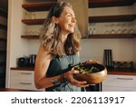 Small photo of Aging woman smiling happily while holding a buddha bowl in her kitchen. Happy senior woman serving herself a healthy vegan meal at home. Mature woman taking care of her body with a plant-based diet.