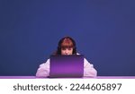 Small photo of Woman with ginger hair looking at the camera while sitting behind her gaming laptop in a studio. Female gamer using an interactive streaming service while sitting in neon purple light.