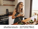 Happy vegan woman smiling while holding a glass of green juice in her kitchen. Mature woman serving herself organic food at home. Woman taking care of her aging body with a healthy plant-based diet.