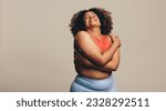 Small photo of Body confident woman embracing her body, expressing self-love and self-acceptance. Young plus-size woman standing in a studio in fitness clothing, embracing her natural physique with joy.