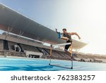 Small photo of Runner jumping over an hurdle during track and field event. Athlete running a hurdle race in a stadium.