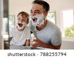Man and little boy with shaving foam on their faces looking into the bathroom mirror and laughing. Father and son having fun while shaving in bathroom.