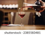 Small photo of Professional female sommelier pours red wine from decanter to the glass, close up image. Woman waiter pouring alcoholic drink being in cellar of wine shop.
