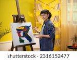 A beautiful painter woman with a very strong jaw is holding a large art palette and painting something on a canvas while admiring her work