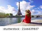 Small photo of Young traveler woman in red dress and hat sitting on the quay of Seine River looking at Eiffel Tower, famous landmark and travel destination in Paris.