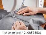 Small photo of Tailor hands stitching gray fabric on modern sewing machine at workplace in atelier. Women's hands sew pieces of fabric on sewing machine closeup. Handmade, hobby, repair, small business concept