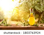 Small photo of Glass container with olive oil on wooden table with branches and olives in crop field full of olive trees with sunshine