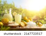 Lemonade with ice in a pitcher and glass on a wooden table with fruit and crushed ice outside with a lemon field in the background on a sunny day. Front view. Horizontal composition.