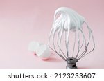 Small photo of Whipped egg whites - beaten italian meringue on a wire whisk and egg shells on pink background.