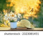 Small photo of summer nature background. chamomile flowers in mug, butterflies, book and braided hat on table in garden. summer season. Beautiful rustic floral composition. relaxation, harmony atmosphere.