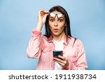 Surprised caucasian young pretty woman looks excitedly at the camera, takes off glasses, holds smartphone, received an unexpected message, saw interesting news, stands on isolated blue background