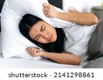 Small photo of Asian young woman, lying in bed at home, covers her ears with a pillow, tries to sleep, closes herself from noise, has a bad dream, restless dream. Sleep and rest concept