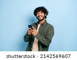 Small photo of Happy handsome amazed stylish indian or arabian guy, using smartphone, chatting online, texting message, browsing internet, social media, looks at camera, stands on isolated blue background, smiling