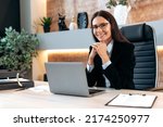 Small photo of Portrait of a gorgeous smart successful caucasian brunette business woman with glasses, company ceo, top manager, sitting at a desk in a modern office, looking at the camera, smiling positively